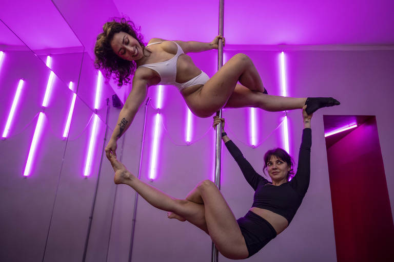 Duas mulheres executam movimentos de pole dance em um estúdio iluminado por luzes roxas verticais. Uma está suspensa na barra com roupa clara, enquanto a outra, com roupa escura, segura a barra e a perna da parceira.