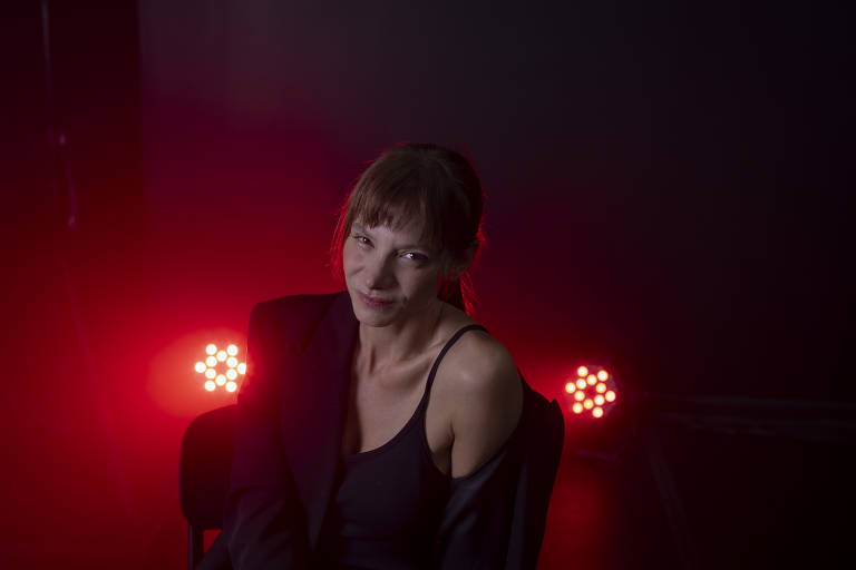 Mulher de cabelos castanhos e franja está sentada, vestindo regata preta e blazer escuro caído no ombro esquerdo. Fundo escuro com duas luzes vermelhas brilhantes atrás dela.