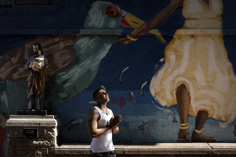 Homem de camiseta regata branca e óculos está em posição de oração diante de estátua de santo segurando criança, com mural colorido ao fundo mostrando figura feminina e pássaro.