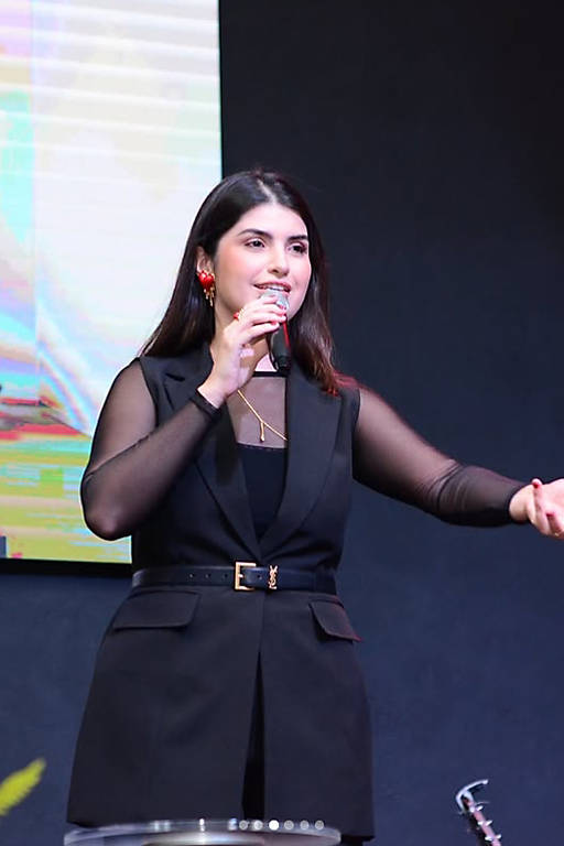 Mulher de cabelos longos e escuros segura microfone com a mão direita e gesticula com a esquerda. Ela veste colete preto com cinto e blusa de manga longa transparente preta, em ambiente interno com fundo escuro e tela colorida ao fundo.