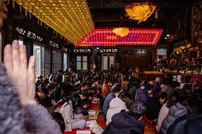 Multidão sentada no chão de templo budista decorado com lanternas amarelas e vermelhas no teto. Pessoa em primeiro plano com mãos em prece, ambiente interno com iluminação suave.